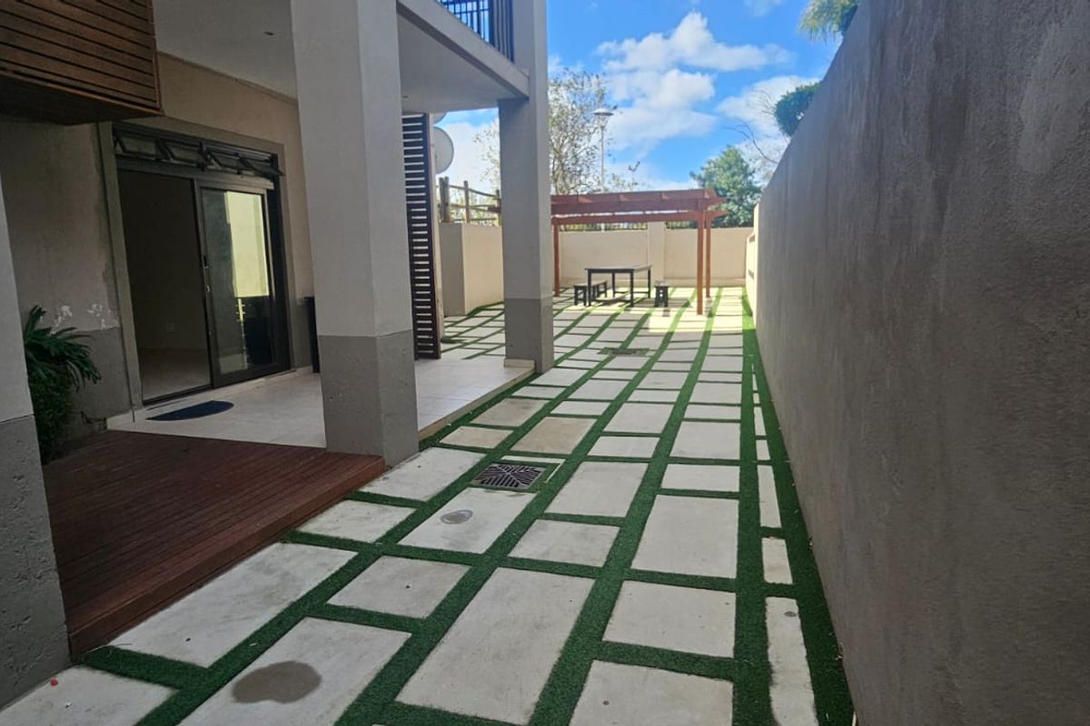 Rear exterior area featuring a covered patio with timber decking, concrete pavers with artificial grass inlay, and a pergola structure.