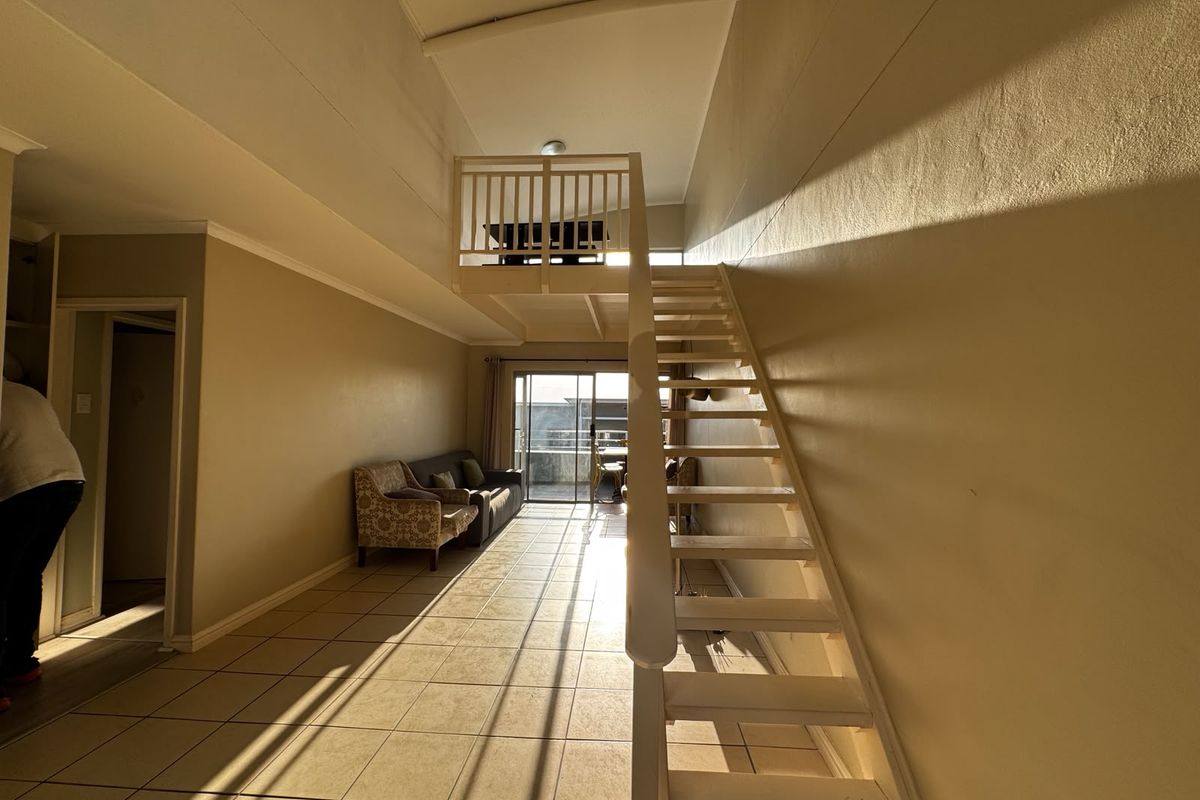 Open-plan living area featuring a mezzanine level with a timber staircase, tiled flooring, and access to a balcony through sliding glass doors.