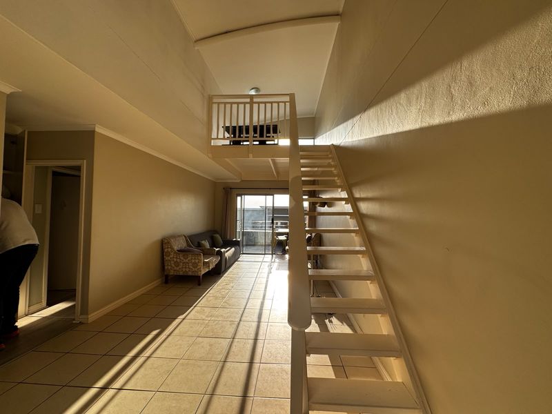 Open-plan living area featuring a mezzanine level with a timber staircase, tiled flooring, and access to a balcony through sliding glass doors.