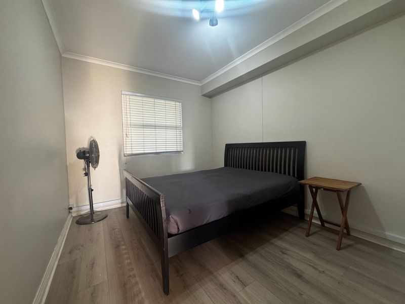A functional bedroom featuring laminate flooring, neutral wall finishes, and a window fitted with venetian blinds.