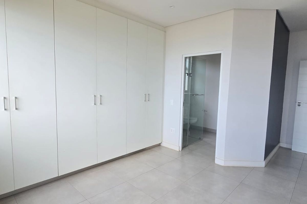 A bright, modern bedroom featuring extensive built-in cupboards, neutral tiled flooring, and direct access to an en-suite bathroom.