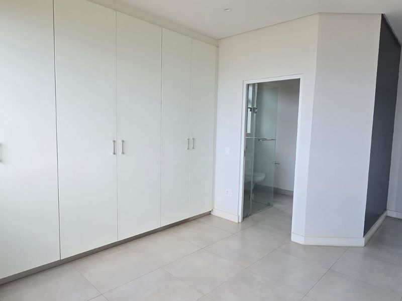 A bright, modern bedroom featuring extensive built-in cupboards, neutral tiled flooring, and direct access to an en-suite bathroom.