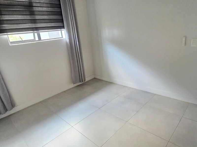 A bright, empty bedroom featuring neutral tiled flooring, clean white walls, and a window fitted with modern blinds.