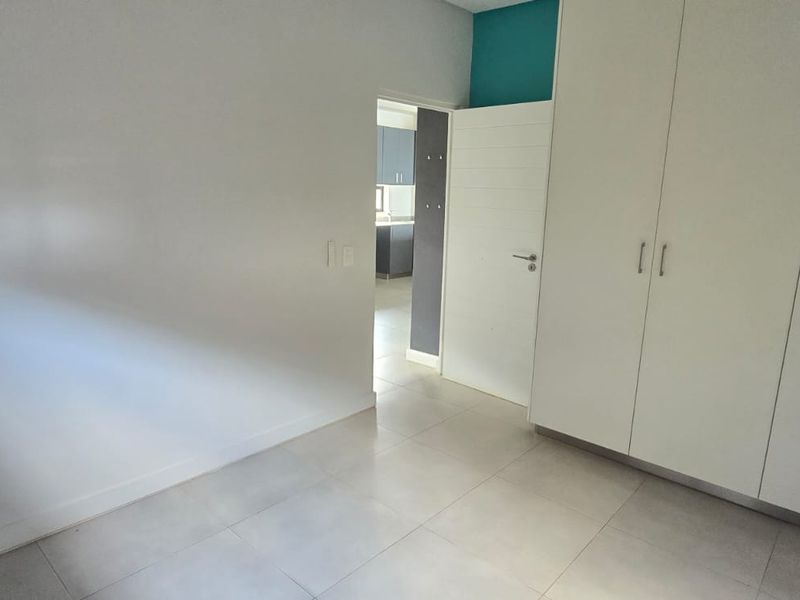 A bright, empty bedroom featuring neutral tiled flooring and ample built-in cupboard storage, with a view through to an adjacent kitchen area.