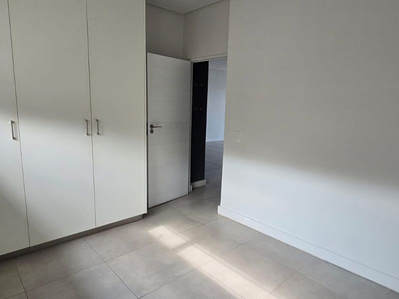 A clean, modern bedroom featuring neutral tiled flooring, built-in cupboards, and roller blinds, offering a bright and versatile living space.