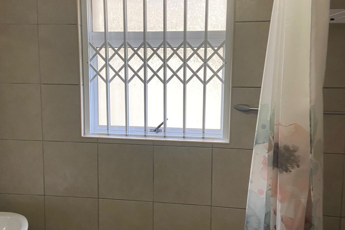 A functional bathroom featuring neutral wall tiles, a pedestal basin, and a window fitted with a security gate.