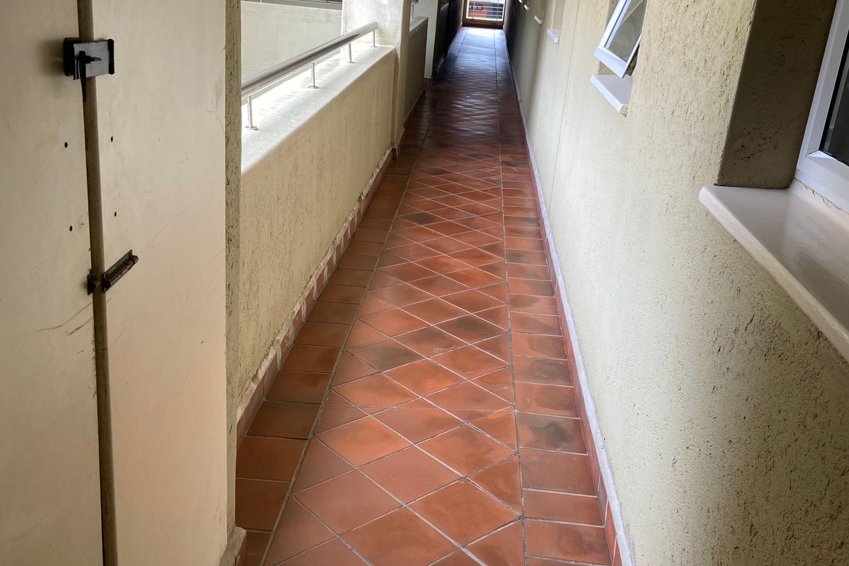 A long, narrow apartment building hallway featuring terracotta floor tiles, metal balustrades, and exterior-facing casement windows.