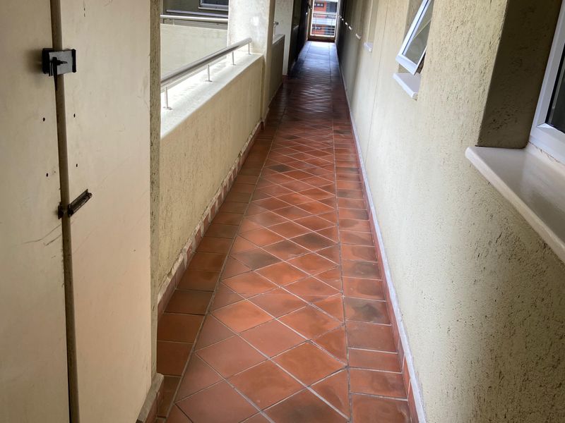 A long, narrow apartment building hallway featuring terracotta floor tiles, metal balustrades, and exterior-facing casement windows.