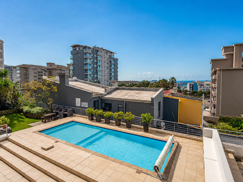 Elevated outdoor pool area featuring a tiled deck, built-in barbecue, and metal safety railings, set against an urban apartment complex backdrop.