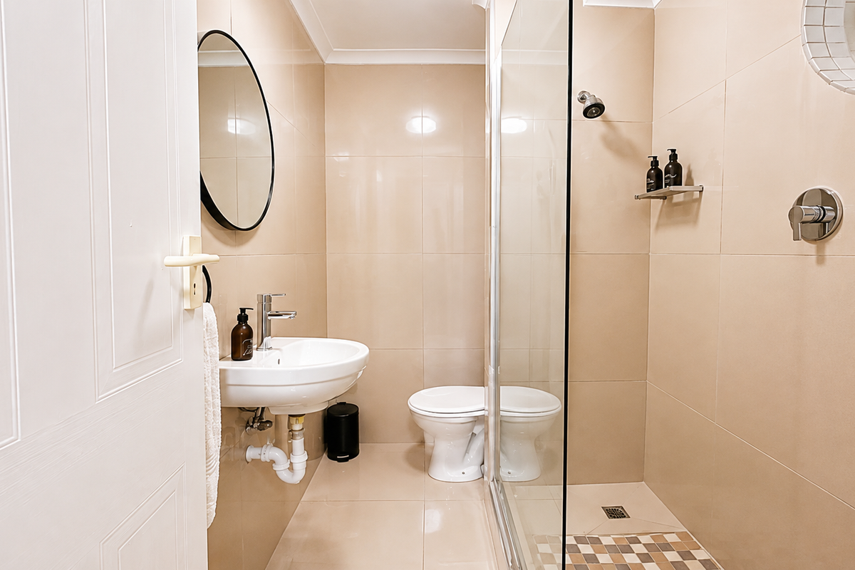 Modern bathroom featuring a glass-enclosed shower, wall-mounted basin, neutral wall tiles, and a unique porthole window, presenting a clean and functional space.
