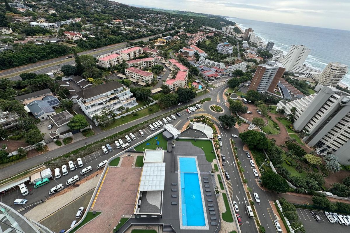 High-angle aerial view of the apartment complex, featuring a central swimming pool, landscaped grounds, and surrounding coastal urban infrastructure.