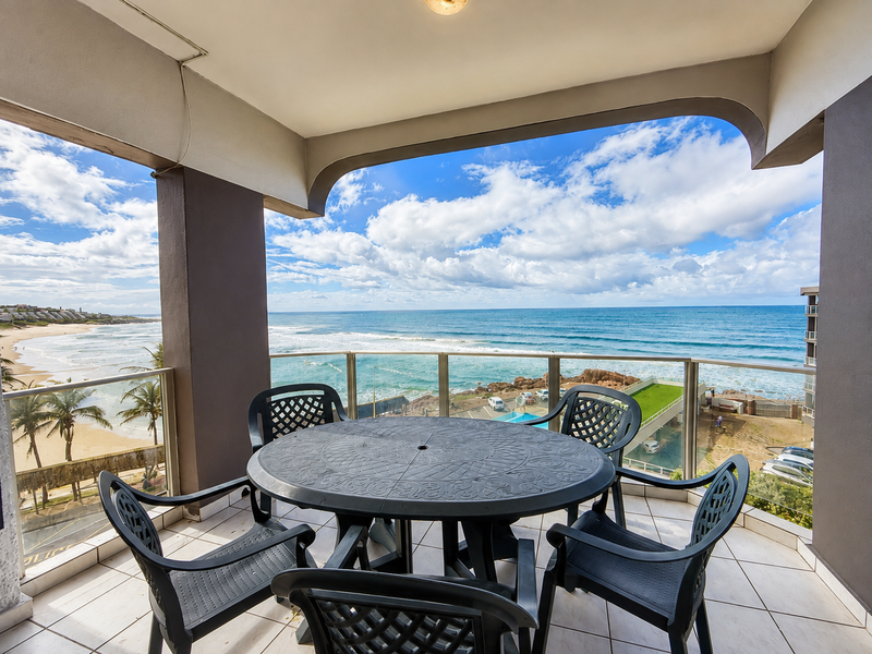 A spacious covered balcony featuring tiled flooring and a glass balustrade, offering panoramic ocean and coastal views.