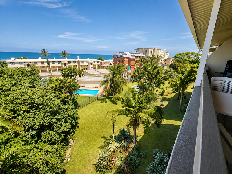 Elevated view from a balcony overlooking a lush garden, swimming pool, and the ocean in the distance.
