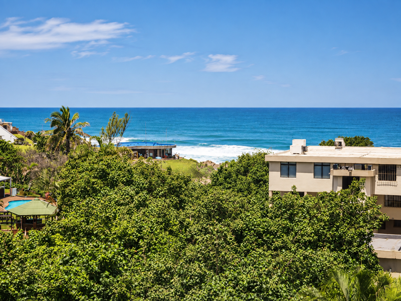 A scenic elevated view overlooking lush coastal greenery, a nearby building, and the expansive blue ocean horizon.