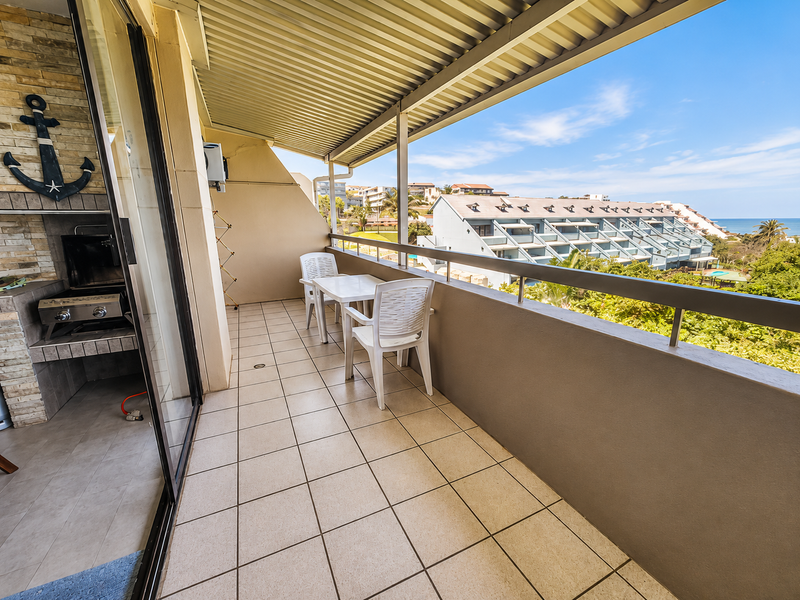 A covered balcony featuring tiled flooring, a built-in braai, and a metal railing, offering pleasant views of the surrounding complex and landscape.