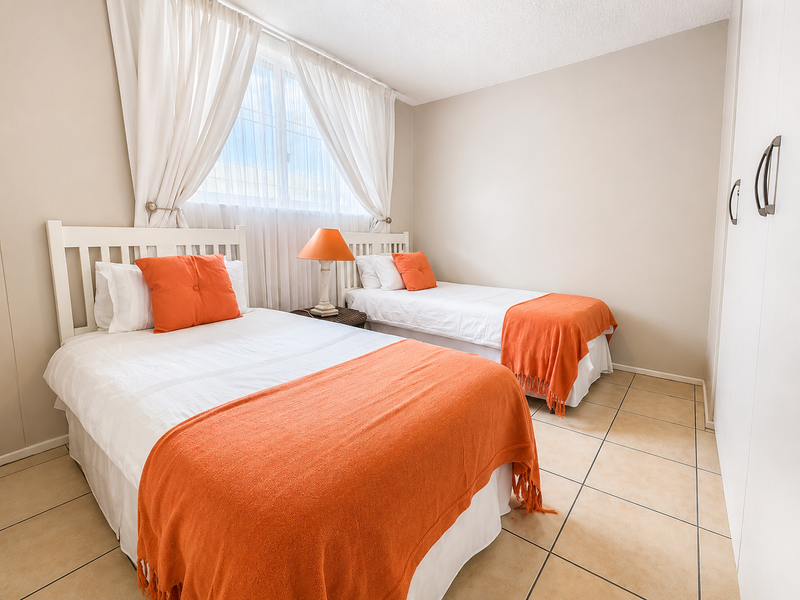 A bright bedroom featuring tiled flooring, a built-in wardrobe, and a window with curtains, comfortably accommodating two single beds.