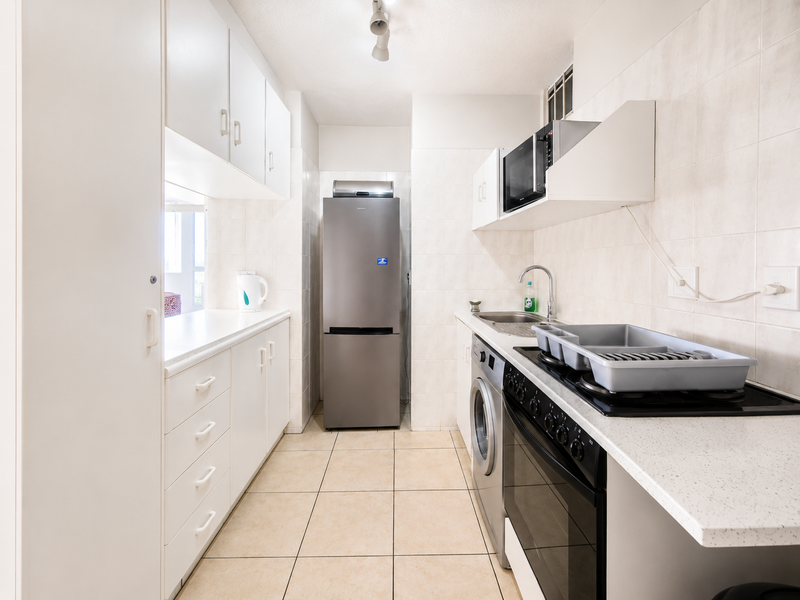 A functional galley-style kitchen featuring white cabinetry, tiled surfaces, a built-in oven and hob, and integrated laundry appliance space.