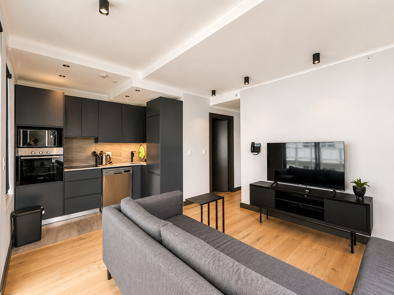 Modern open-plan living area featuring a sleek kitchen with dark cabinetry, integrated appliances, timber flooring, and recessed lighting, creating a contemporary and functional space.