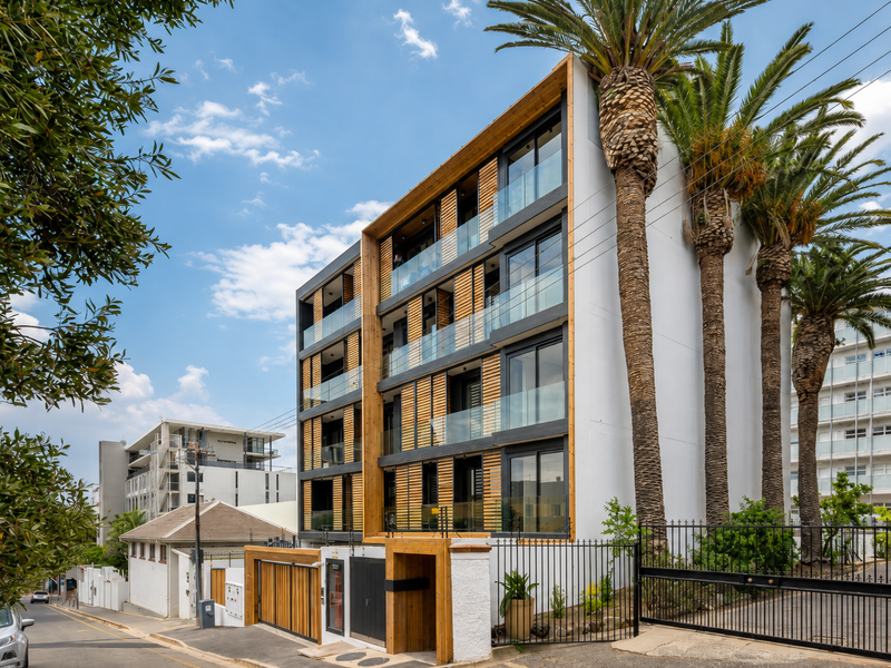 Modern apartment building exterior featuring timber cladding, glass balconies, and secure gated access with mature palm trees.