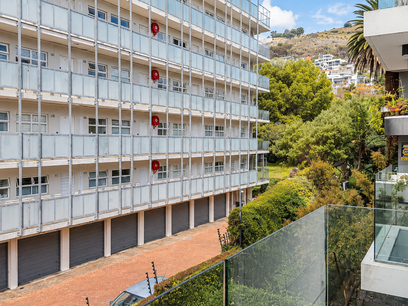 An elevated view overlooking the apartment complex exterior, featuring glass balustrades, ground-level garage access, and surrounding greenery.