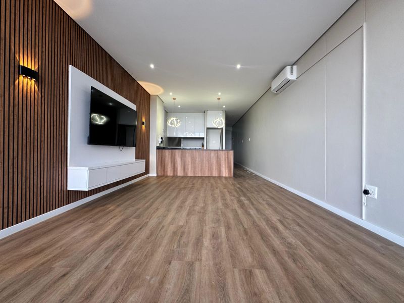Modern open-plan living area featuring timber slat wall detailing, built-in media console, laminate flooring, and a sleek kitchen with island and pendant lighting.