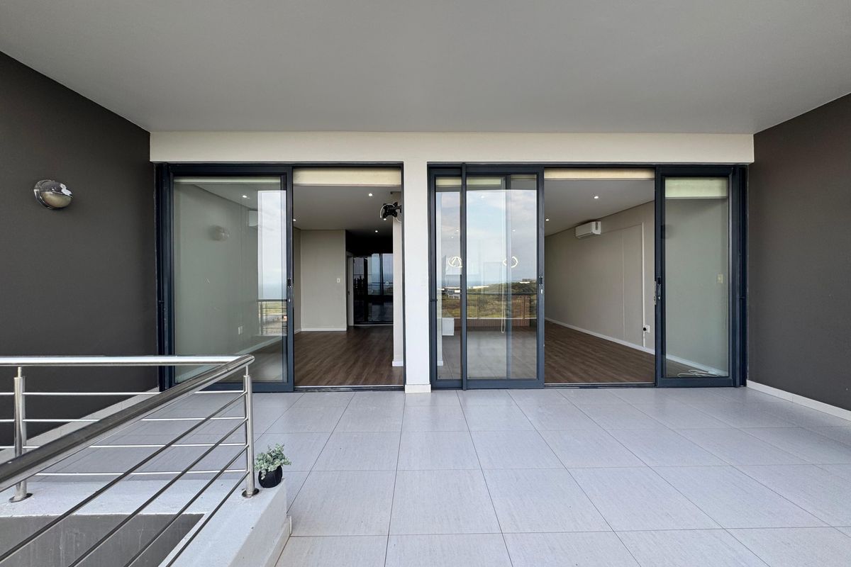 A spacious, modern covered balcony featuring large sliding glass doors, sleek stainless steel balustrades, and neutral floor tiling with recessed lighting.