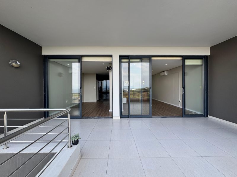 A spacious, modern covered balcony featuring large sliding glass doors, sleek stainless steel balustrades, and neutral floor tiling with recessed lighting.