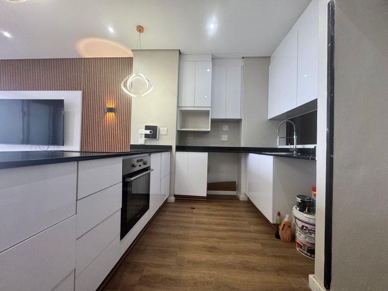 Modern kitchen featuring white gloss cabinetry, black granite countertops, built-in oven, and wood-look flooring, adjacent to a stylish slatted wood feature wall.