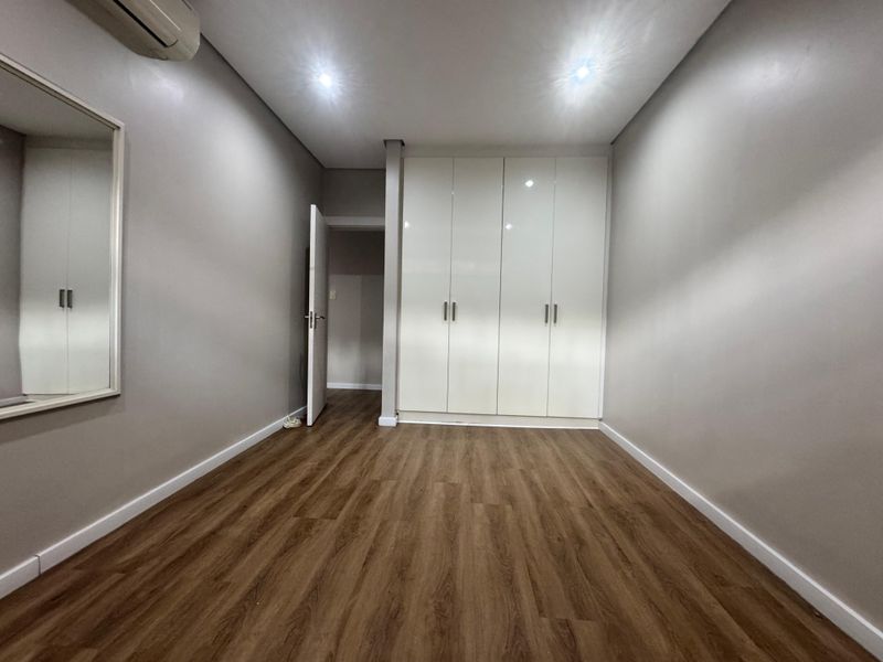A clean, empty bedroom featuring modern laminate flooring, built-in white cupboards, recessed lighting, and a wall-mounted air conditioning unit.