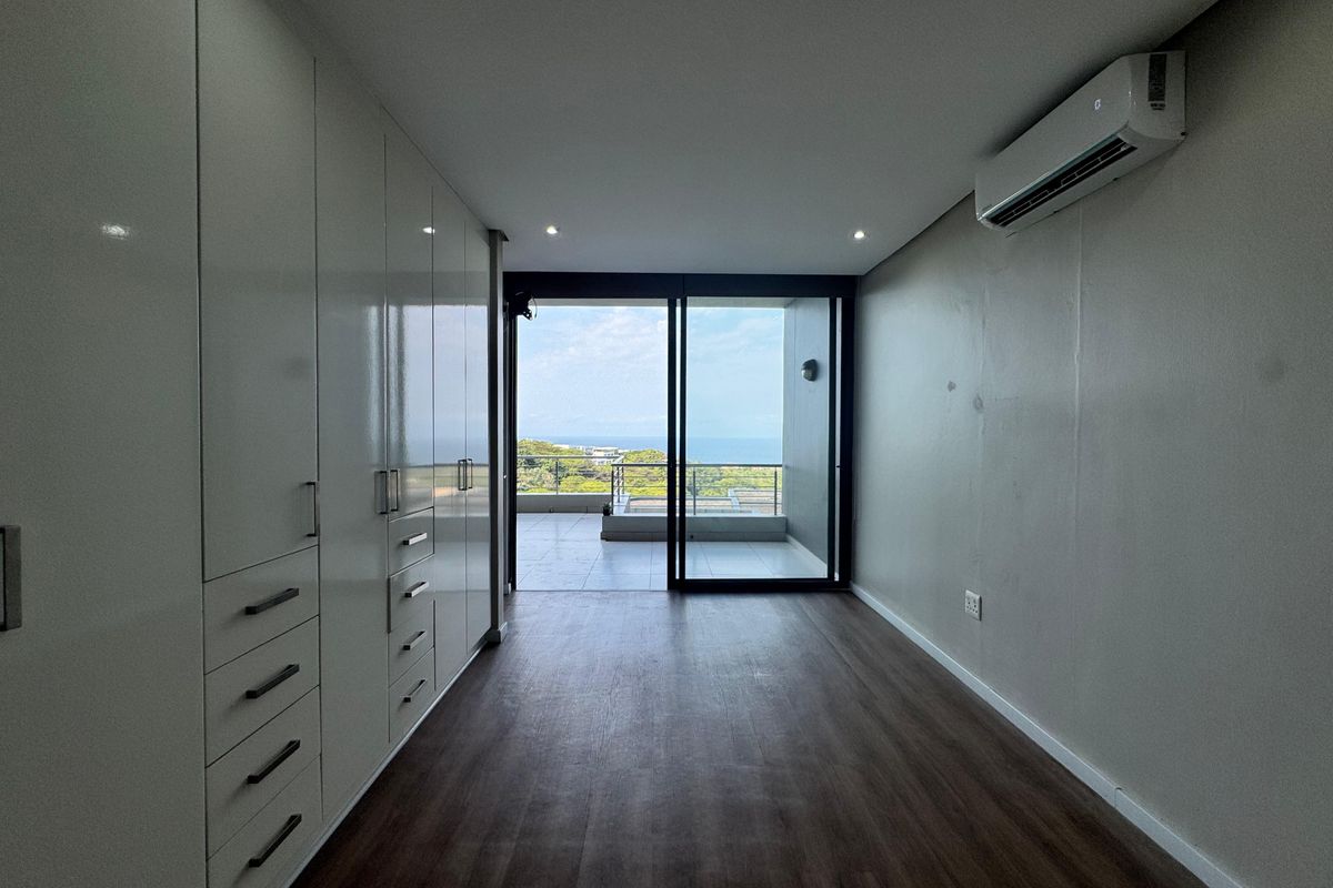 A bright, empty bedroom featuring extensive built-in cabinetry, modern laminate flooring, a split-system air conditioner, and direct access to a private balcony with ocean views.