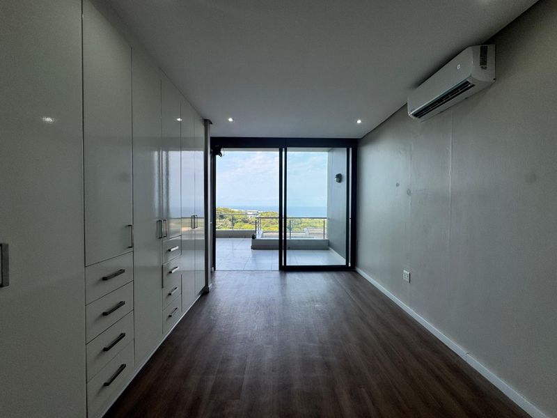 A bright, empty bedroom featuring extensive built-in cabinetry, modern laminate flooring, a split-system air conditioner, and direct access to a private balcony with ocean views.