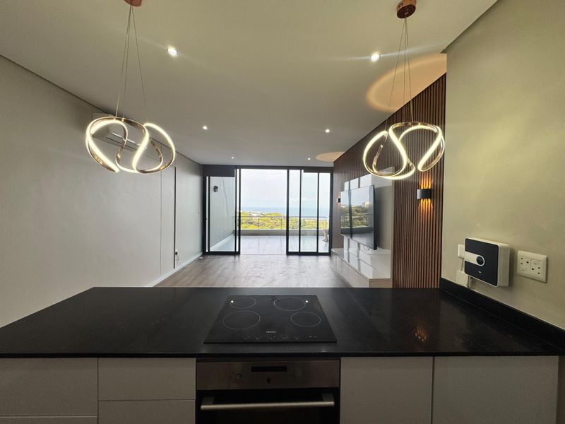 Modern open-plan kitchen and living area featuring a sleek built-in oven, electric hob, stone countertops, and direct access to a balcony with scenic views.