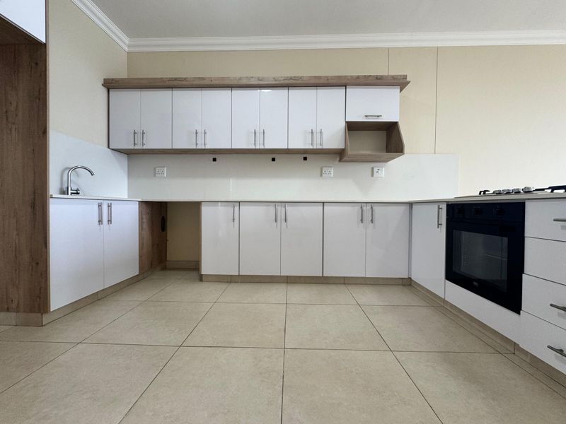 A modern, clean kitchen featuring white cabinetry with wood-tone accents, built-in oven, gas hob, and neutral tiled flooring in an open-plan layout.