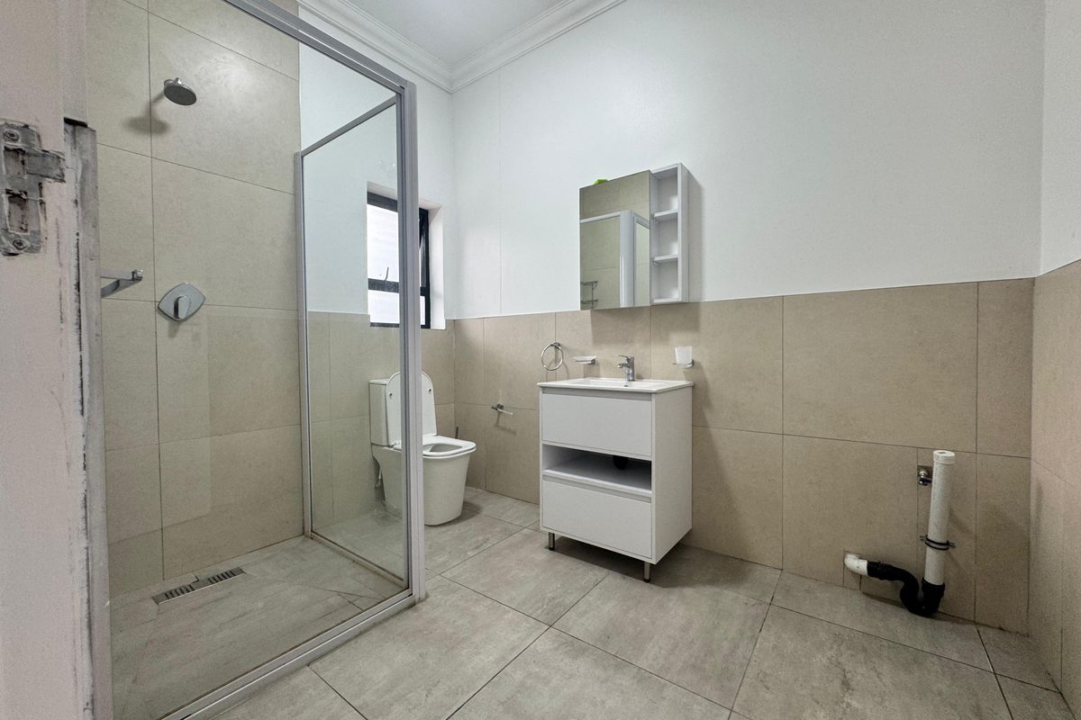 A modern bathroom featuring a glass-enclosed shower, vanity unit with basin, and neutral wall and floor tiling. Requires finishing touches to plumbing and door frame.