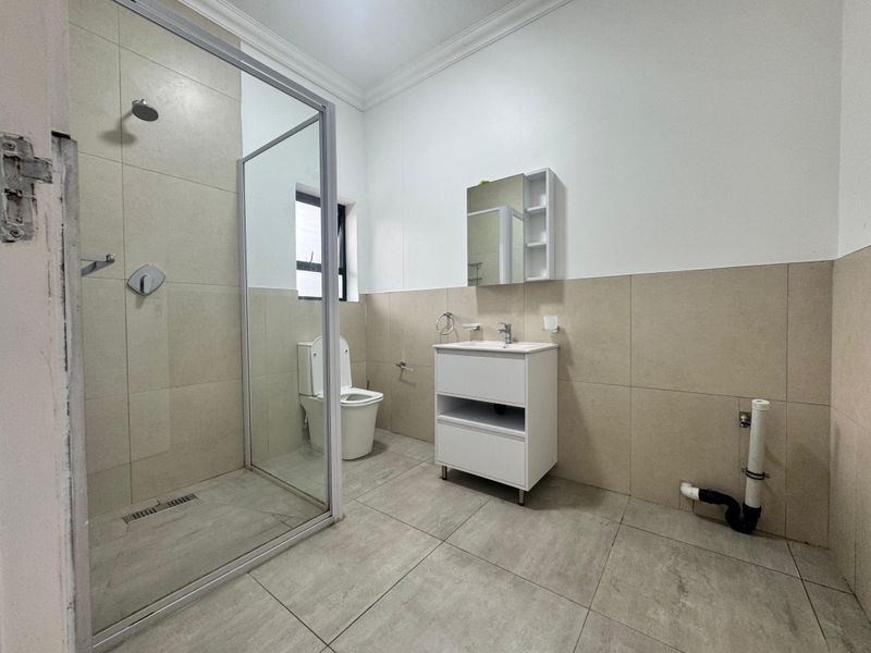 A modern bathroom featuring a glass-enclosed shower, vanity unit with basin, and neutral wall and floor tiling. Requires finishing touches to plumbing and door frame.
