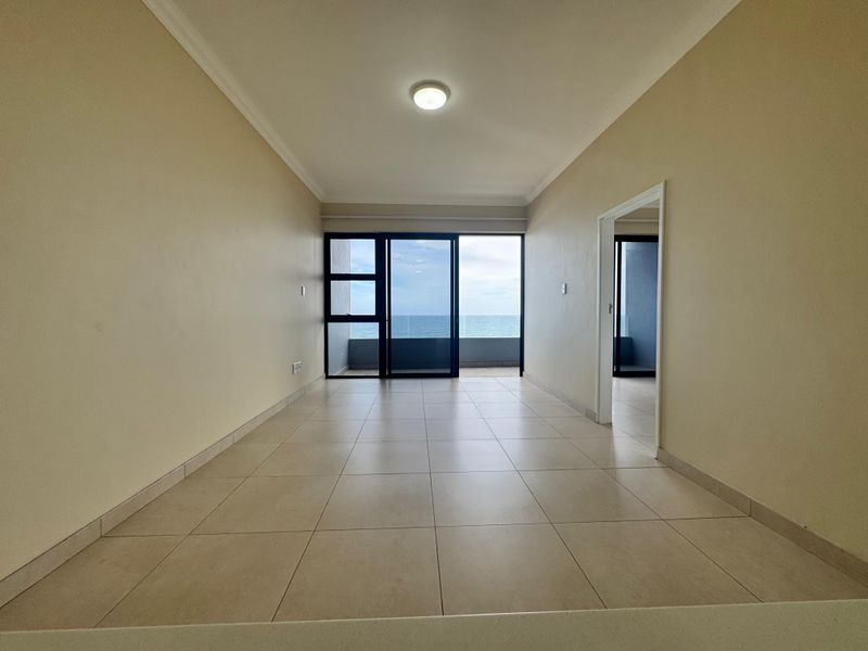 An empty, open-plan living area featuring neutral tiled flooring, coving, and a large sliding glass door providing access to a balcony with ocean views.