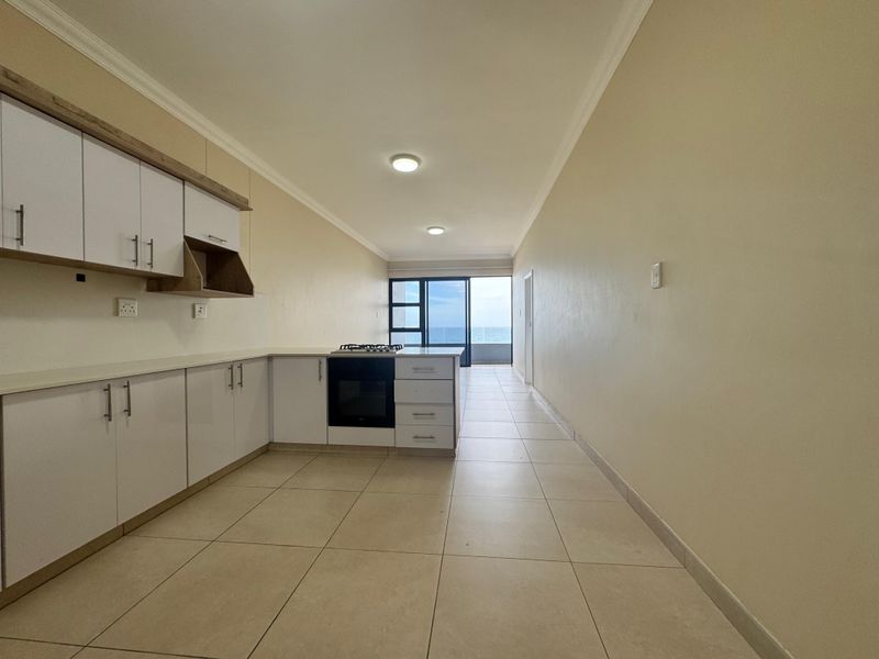Open-plan kitchen and living area featuring modern white cabinetry, built-in oven and hob, tiled flooring, and direct access to a balcony with ocean views.