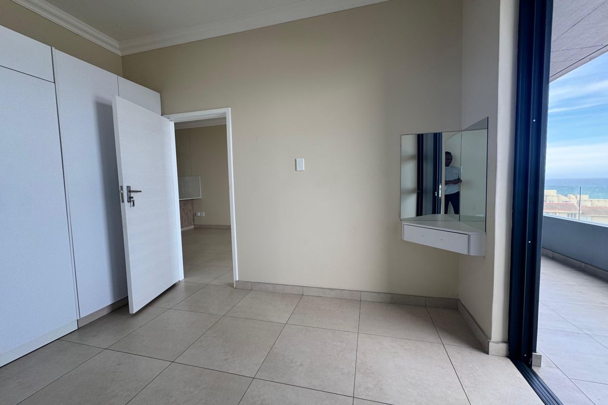 A bright, empty bedroom featuring tiled floors, built-in cupboards, a floating vanity, and direct access to a balcony with ocean views.