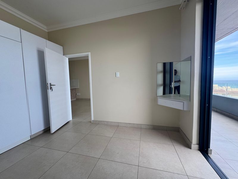A bright, empty bedroom featuring tiled floors, built-in cupboards, a floating vanity, and direct access to a balcony with ocean views.