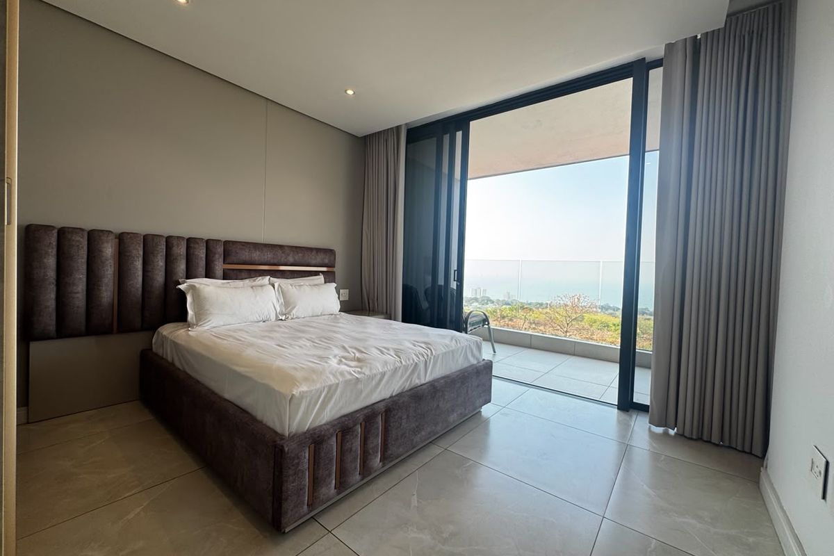Modern bedroom featuring large format floor tiles, recessed lighting, and expansive sliding glass doors opening onto a balcony with scenic coastal views.
