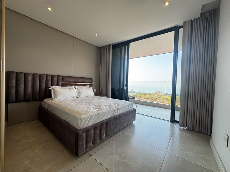 Modern bedroom featuring large format floor tiles, recessed lighting, and expansive sliding glass doors opening onto a balcony with scenic coastal views.