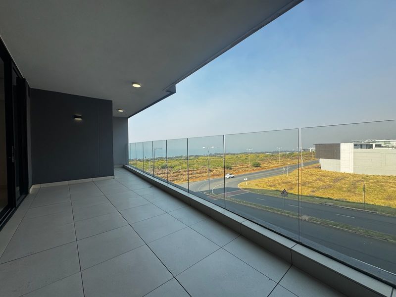 A spacious, modern balcony featuring a frameless glass balustrade, tiled flooring, and recessed lighting, offering an expansive view of the surrounding landscape.