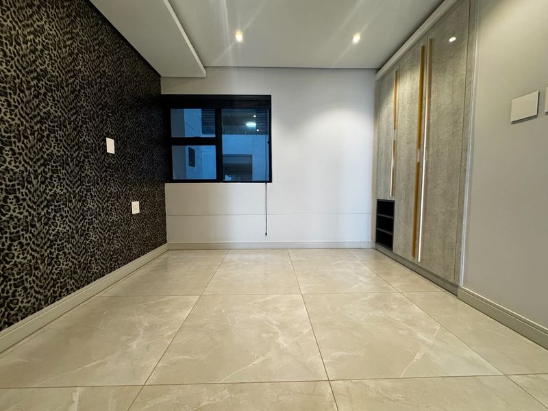 A bright, empty bedroom featuring large format floor tiles, modern built-in cupboards with integrated lighting, and a feature wall.