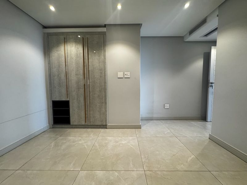 A clean, modern bedroom featuring neutral porcelain floor tiles, recessed ceiling lighting, and a contemporary built-in wardrobe with integrated shelving.