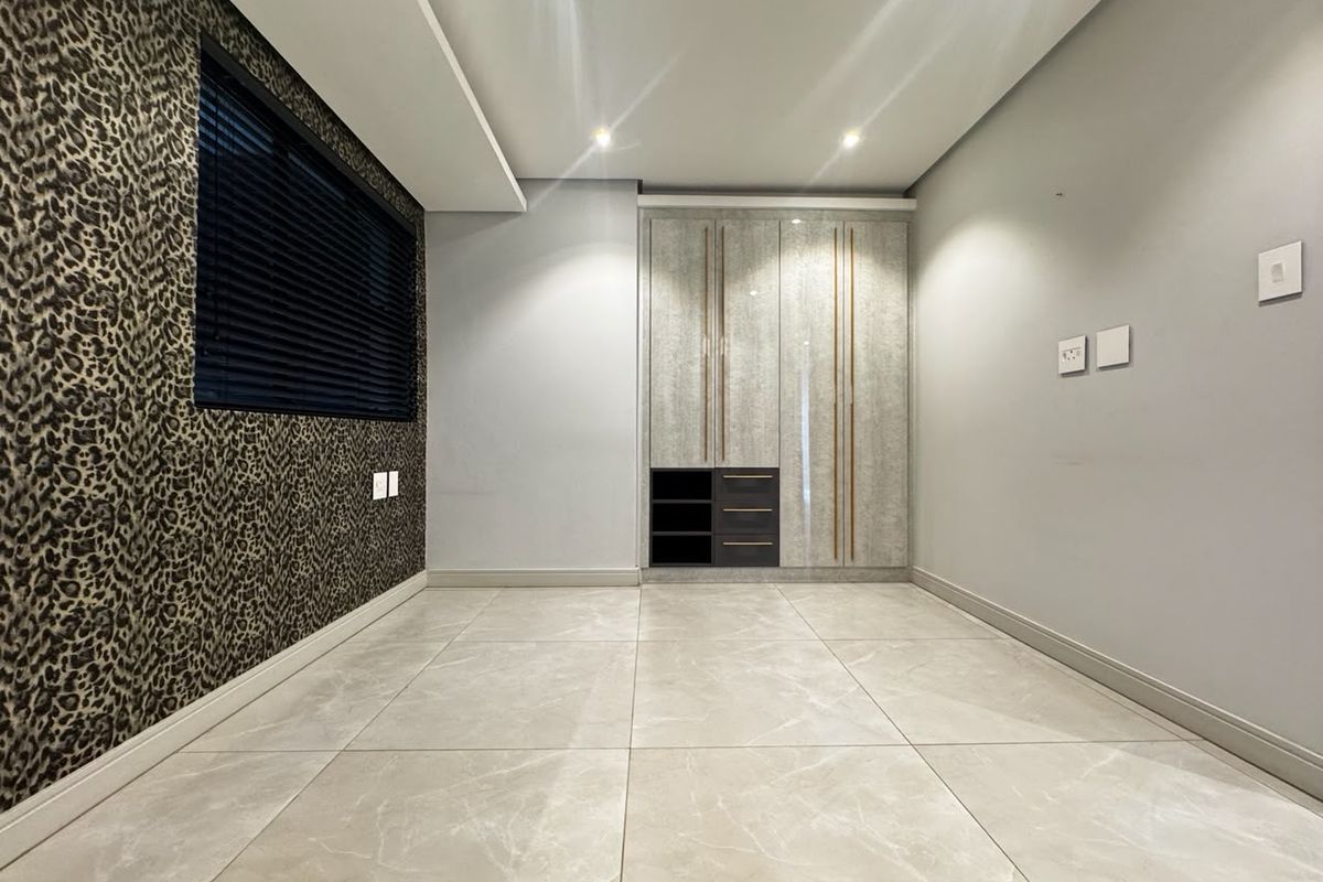 A modern, empty bedroom featuring large-format porcelain floor tiles, recessed ceiling lighting, and a sleek, built-in wardrobe with integrated shelving.