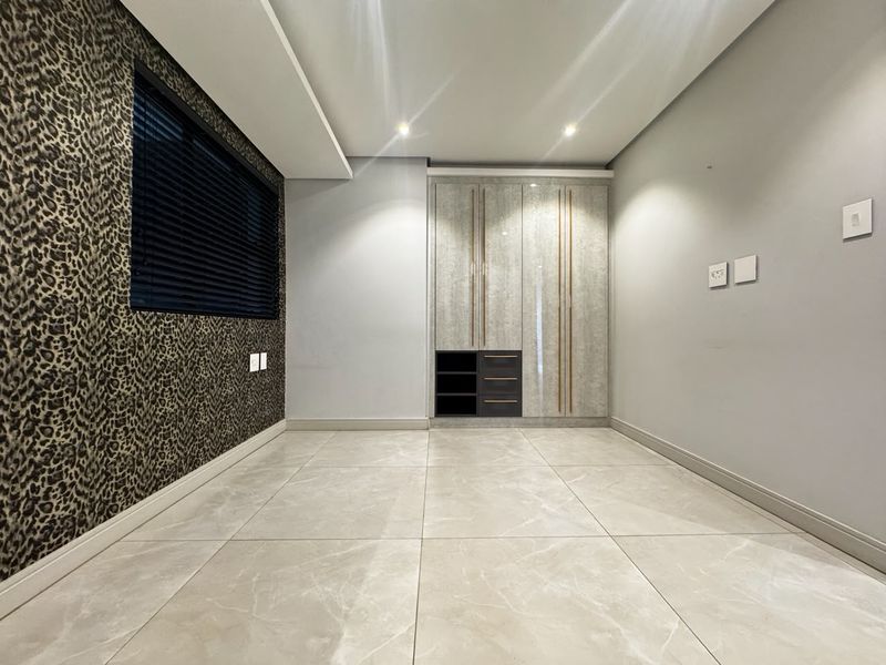 A modern, empty bedroom featuring large-format porcelain floor tiles, recessed ceiling lighting, and a sleek, built-in wardrobe with integrated shelving.