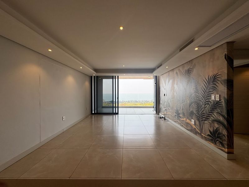 Spacious, empty living area featuring large format floor tiles, recessed lighting, and a decorative feature wall, opening onto a balcony with scenic views.