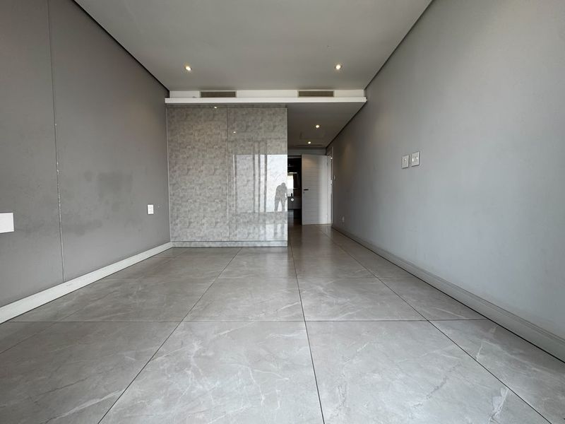 A spacious, empty living area featuring modern large-format porcelain floor tiles, recessed downlighting, ducted air conditioning, and sleek built-in cabinetry.
