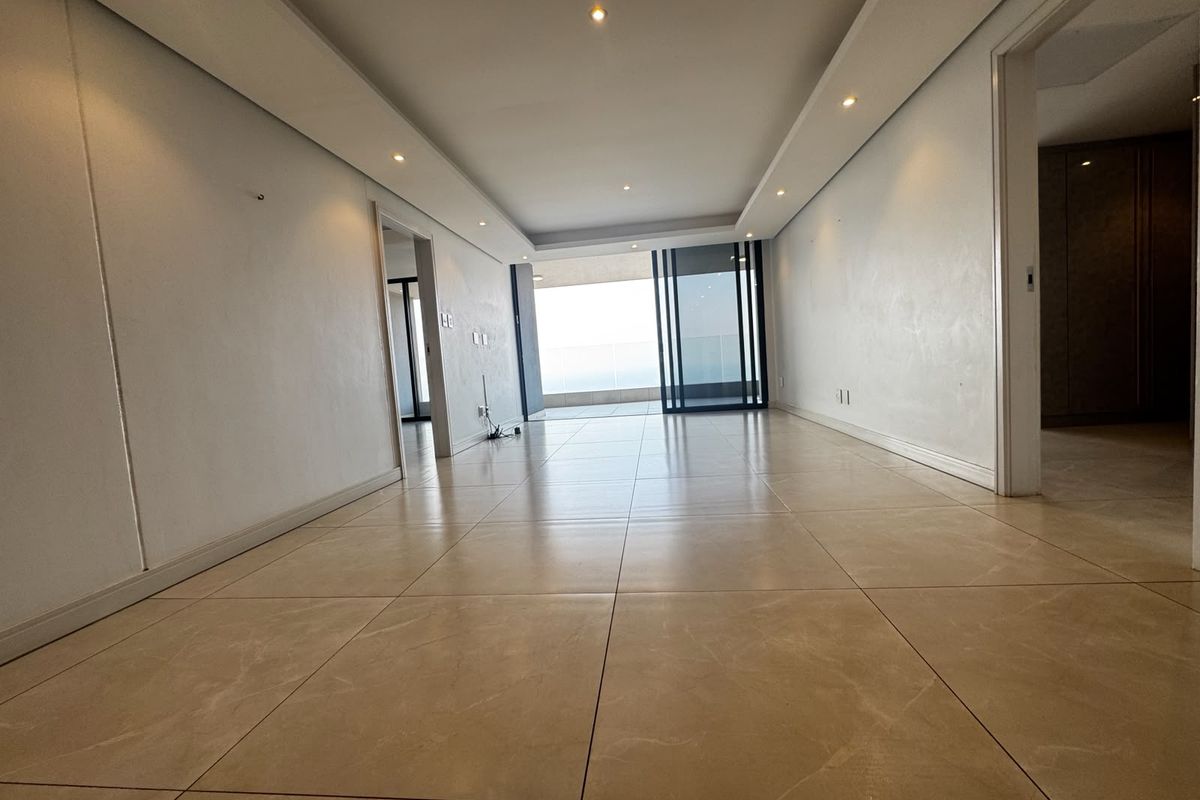 A spacious, empty living area featuring neutral tiled flooring, recessed ceiling lighting, and a large sliding glass door leading to an outdoor balcony.