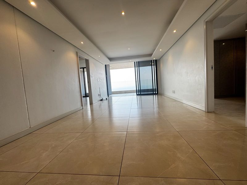 A spacious, empty living area featuring neutral tiled flooring, recessed ceiling lighting, and a large sliding glass door leading to an outdoor balcony.